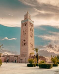 Stunning view of the Koutoubia Mosque in Marrakech, Morocco, with its towering minaret set against a dramatic sky. The surrounding plaza is quiet, adorned with palm trees and well-kept greenery, capturing the serene beauty of this historic landmark.